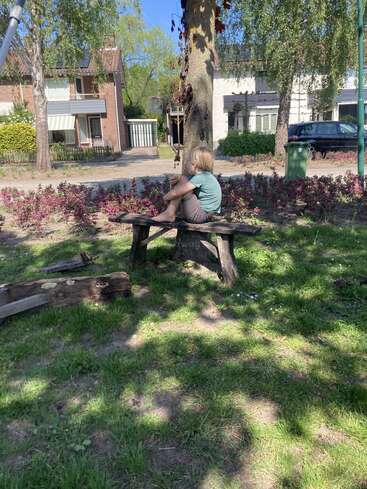 Un niño pequeño está sentado descalzo en un banco de madera al aire libre, rodeado de vegetación y árboles, con la mirada fija en el horizonte. Al fondo se ven casas y coches de las afueras.