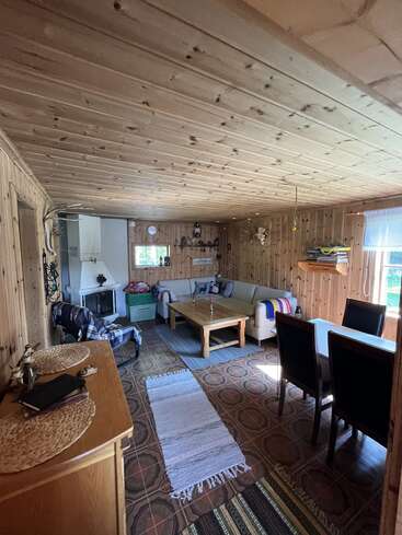 This cozy wooden cabin living room features a corner fireplace, comfortable sofa, coffee table, dining area, books, rugs, bright window, and rustic decor elements throughout.