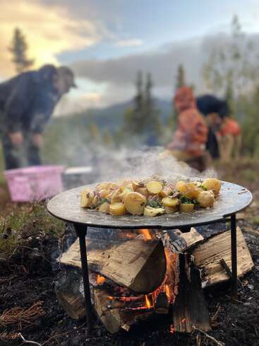 Golden potatoes cook on a flat grill over an open fire outdoors. Two blurred people are in the background, surrounded by nature and trees at sunset.