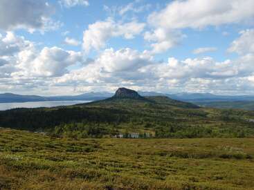 The image depicts a serene landscape featuring a lush green field, a small lake, and a majestic mountain range under a blue sky with white clouds.