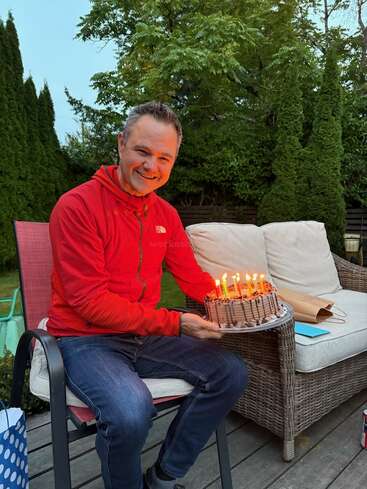 A smiling man in a red jacket sits outdoors, holding a chocolate birthday cake with lit candles. He is celebrating, surrounded by greenery and patio furniture.