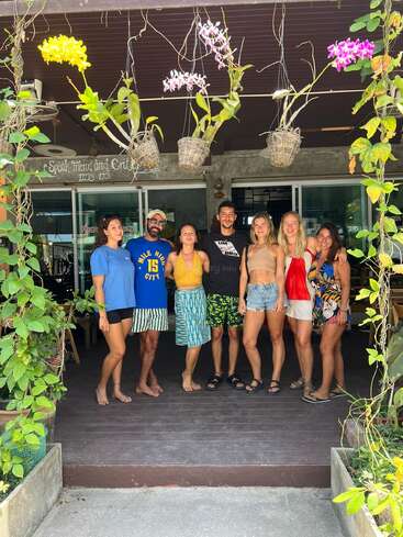 Seven friends are posing together on a patio, surrounded by green plants and hanging orchids. They're barefoot, smiling, and enjoying a sunny, relaxed atmosphere.