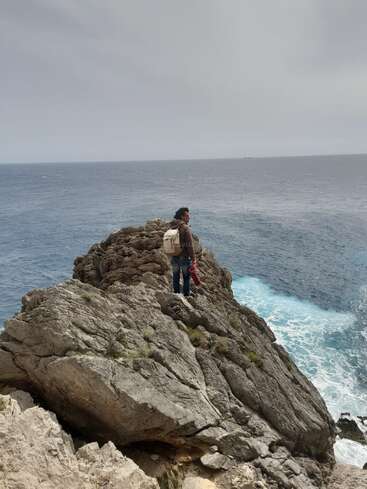 Una persona está de pie en un acantilado rocoso sobre el océano, contemplando el vasto mar bajo un cielo nublado, con una mochila y una chaqueta en la mano.