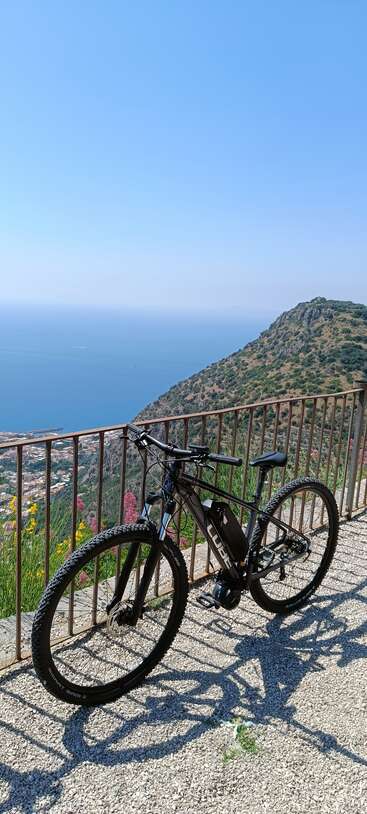 Una bicicleta de montaña negra se apoya en una barandilla metálica, con vistas a una ciudad costera, una colina y un vasto mar azul bajo un cielo claro y brillante. Un paisaje sereno.