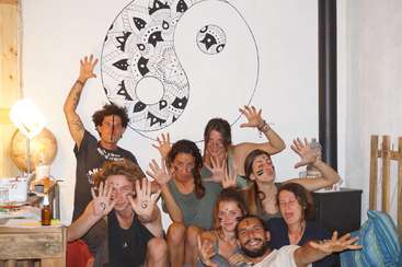 A imagem mostra um grupo de pessoas reunidas, algumas sentadas no chão e outras em pé, todas de frente para a câmera, em uma sala com mesa e cadeiras.