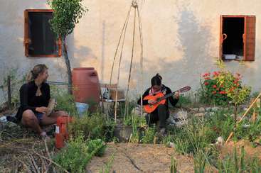 Duas pessoas estão tocando instrumentos de corda em um jardim, cercado por uma cerca, com uma mesa e cadeiras ao fundo, criando uma atmosfera pacífica e serena.