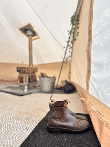 Um par de botas marrons gastas está sobre um tapete dentro de uma barraca aconchegante. Um fogão a lenha arde ao fundo, criando um ambiente acolhedor.