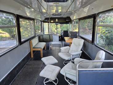 Esta imagem mostra o interior de um ônibus reformado, com poltronas confortáveis, um banco, uma mesa, janelas amplas e um ambiente limpo e minimalista.