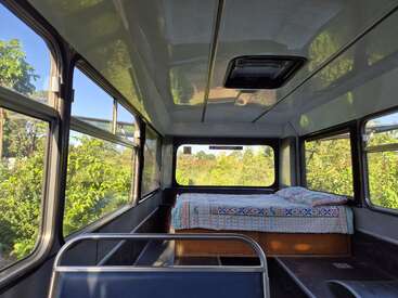 Esta imagem mostra o interior de um ônibus transformado em um espaço habitável, com uma cama bem arrumada coberta por uma colcha estampada, rodeada por janelas amplas.