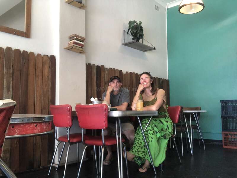 Um homem e uma mulher sentados em uma mesa de café retrô com cadeiras vermelhas, sorrindo. A sala tem paredes azul-petróleo, painéis de madeira e prateleiras com livros e plantas.