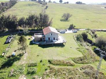 Aerial view of a solitary house with a red roof, surrounded by fields, trees, cars, and open green space in a peaceful rural landscape.
