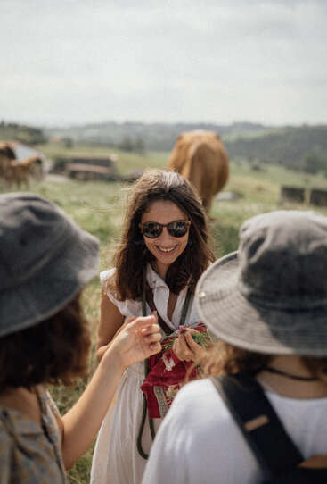 Three women enjoy a sunny day outdoors in the countryside, smiling and chatting, with cows grazing in the background. Two wear hats, one sunglasses.