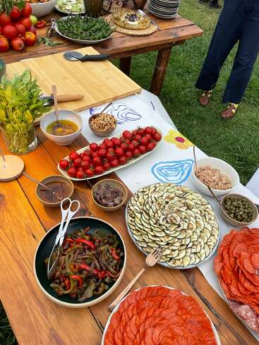 An outdoor buffet displays sliced vegetables, fresh tomatoes, herbs, charcuterie, dressings, nuts, capers, and bread on wooden tables, suggesting a vibrant, rustic picnic setting.