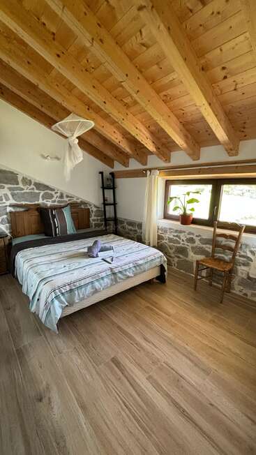 This cozy bedroom features a wooden beamed ceiling, stone walls, a neatly made bed, wooden furniture, large window with plant, and light-filled serene atmosphere.