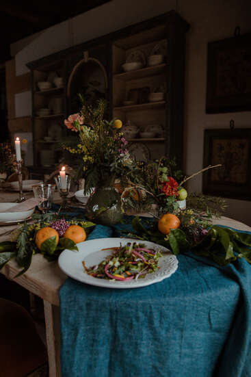 A rustic dinner table decorated with candles, vibrant flowers, and fresh oranges. A blue tablecloth and a plate of salad create a cozy, inviting atmosphere.