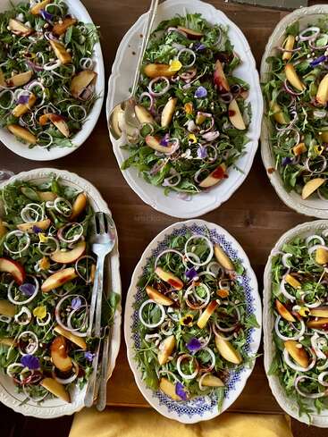 Six oval platters of fresh salad on a wooden table, featuring greens, peach slices, red onion rings, edible flowers, and pine nuts, beautifully arranged.