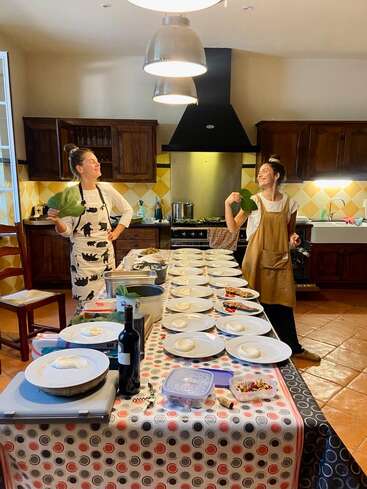 Two women in aprons stand in a kitchen, smiling and holding leaves, with a long table set with plates, wine, and prepared ingredients ready to serve.