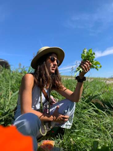 A woman wearing sunglasses and a hat squats in tall grass on a sunny day, holding fresh greens, possibly foraging or gardening, under a clear blue sky.