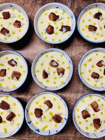 Nine small bowls of creamy soup, garnished with golden croutons, green oil drops, and delicate white edible flowers, arranged artfully on a rustic wooden surface.