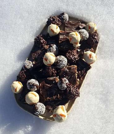 A rectangular platter rests on snow, filled with assorted chocolate treats: dark chocolate squares, coconut-covered balls, nut-coated bites, and white chocolate truffles with almond slivers.