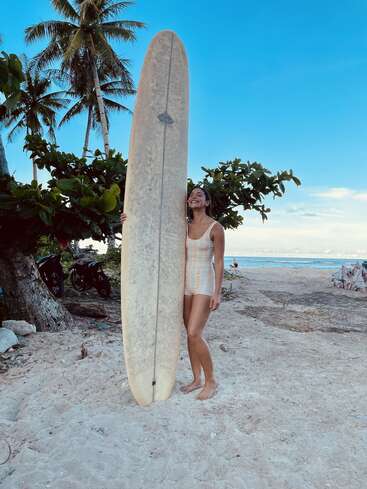 A smiling woman in a swimsuit stands on a sandy beach, holding a tall surfboard. Palm trees and ocean view create a tropical, relaxed atmosphere.