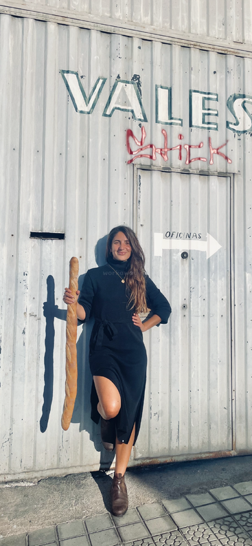 A woman in a black dress stands confidently against a metal wall, holding an oversized baguette. Sunlight casts shadows, and graffiti decorates the industrial background.