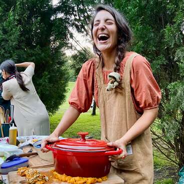 A woman wearing a brown apron and orange blouse laughs joyfully outdoors, standing behind a red pot on a table with food, surrounded by greenery and trees.