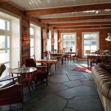 This cozy, sunlit room features large windows, wooden beams, vintage furniture, red velvet chairs, small tables, wall clocks, crystal lamps, and a welcoming rustic ambiance.