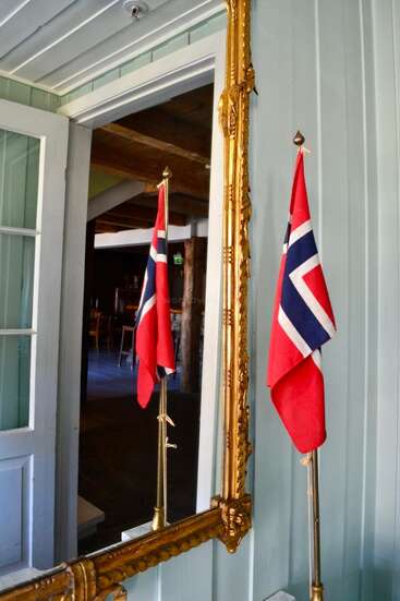 Eine kleine norwegische Flagge steht neben einem goldenen, verzierten Spiegel, dessen Spiegelbild man gut sehen kann. Hellblaue Holzwände sorgen für eine gemütliche, traditionelle skandinavische Innenausstattung.
