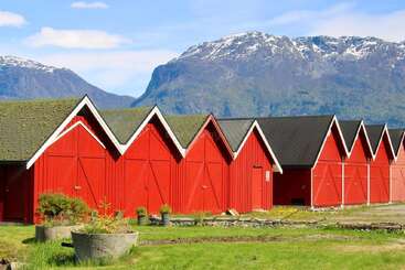 Eine Reihe knallroter Holzscheunen mit spitzen Dächern steht vor dem Hintergrund hoher, schneebedeckter Berge und einer grünen Landschaft unter einem teilweise bewölkten Himmel.