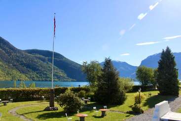 Ein strahlender, sonniger Tag in einem ruhigen Garten an einem See, umgeben von Bergen. Ein Fahnenmast steht hoch, Stühle und Tische sind überall verteilt.