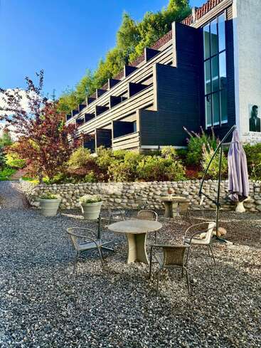 Ein modernes Gebäude mit gestuften Balkonen blickt auf eine Terrasse mit Kiesboden, Tischen, Stühlen, großen Blumentöpfen, Bäumen, einer Steinmauer und leuchtendem Grün unter einem strahlend blauen Himmel.