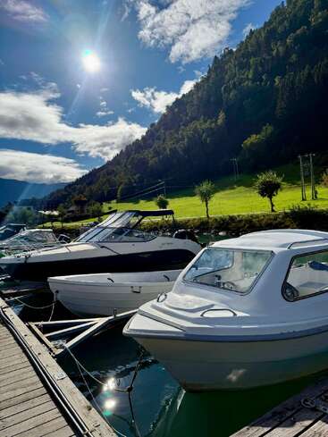Die Boote liegen an einem ruhigen Gewässer neben einem Holzsteg. Grüne Hügel, Bäume und strahlender Sonnenschein unter vereinzelten Wolken sorgen für eine friedliche, malerische und einladende Atmosphäre.