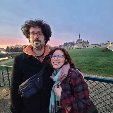 Ein lächelndes Paar steht bei Sonnenuntergang draußen in der Nähe eines Zauns, im Hintergrund ein historisches Schloss, und ein sanfter, farbenfroher Abendhimmel taucht die Szene in Licht.
