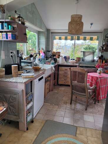This cozy kitchen features vintage furniture, large windows with garden views, a round table with a red cloth, eclectic decor, and various appliances, creating a lived-in charm.