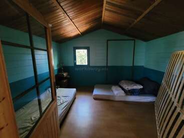 Um quarto simples de madeira com dois colchões no chão, paredes azul-claro, uma janela pequena, algumas roupas de cama e um teto de madeira, com pouquíssimos móveis.
