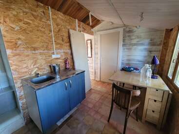 Este quarto aconchegante tem uma pequena cozinha compacta com pia, uma mesa de jantar rústica com um abajur, paredes de madeira, piso de azulejo e móveis simples e funcionais.