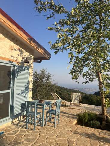 A peaceful patio with light blue wooden chairs and table, stone flooring, leafy tree, summer sunlight, scenic mountain view, and distant blue sea under clear sky.