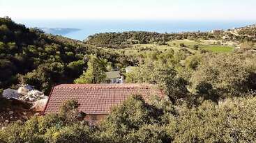 A red-roofed house nestles among dense green trees on a hillside, overlooking fields, distant hills, and the deep blue sea under a clear sky.