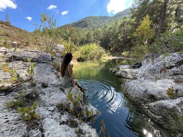 A woman sits on a rocky ledge, dipping her feet into a tranquil green pond surrounded by trees and mountains under a bright, blue sky. Peaceful nature.