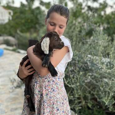 Une jeune fille vêtue d'une robe à fleurs serre doucement une petite chèvre brune dans ses bras. Elles sont en plein air, entourées d'arbres verts, créant un moment de paix et d'affection.