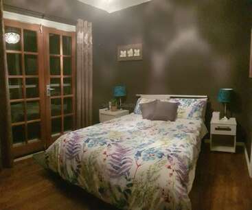 Ein gemütliches Schlafzimmer mit dunklen Wänden, geblümter Tagesdecke, zwei tealfarbenen Lampen, weißen Nachttischen, Flügeltüren, Holzboden, Vorhängen und einem kleinen gerahmten Kunstwerk über dem Bett.