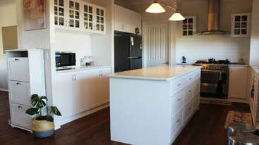 Diese moderne Küche hat weiße Schränke, eine große Mittelinsel, Edelstahlgeräte, Holzböden, viel Stauraum, eine Topfpflanze, Hängeleuchten und eine gemütliche, einladende Atmosphäre.