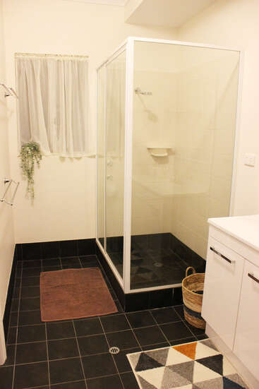 Dieses Badezimmer hat eine Glasdusche, einen schwarz gefliesten Boden, einen weißen Waschtisch, einen dekorativen Teppich, einen Wäschekorb, eine braune Badematte, ein Fenster mit durchsichtigem Vorhang und hängende Grünpflanzen.