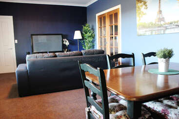 In diesem gemütlichen Wohn- und Esszimmer stehen ein dunkles Sofa, ein Fernseher, ein Esstisch aus Holz, grüne Stühle, Topfpflanzen, blaue Wände und ein Eiffelturm-Kunstwerk.