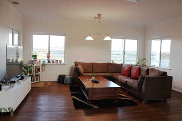 Ein geräumiges Wohnzimmer mit einem großen braunen Sektionssofa, einem Couchtisch aus Holz, einem modernen TV-Ständer, vielen Fenstern, Pflanzen, einem Bücherregal und einer gemütlichen Beleuchtung.