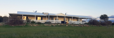 Ein einstöckiges Haus mit einer großen Veranda und weißem Geländer steht auf einer Wiese, umgeben von Felsen, Büschen und dem offenen Himmel bei Sonnenuntergang.