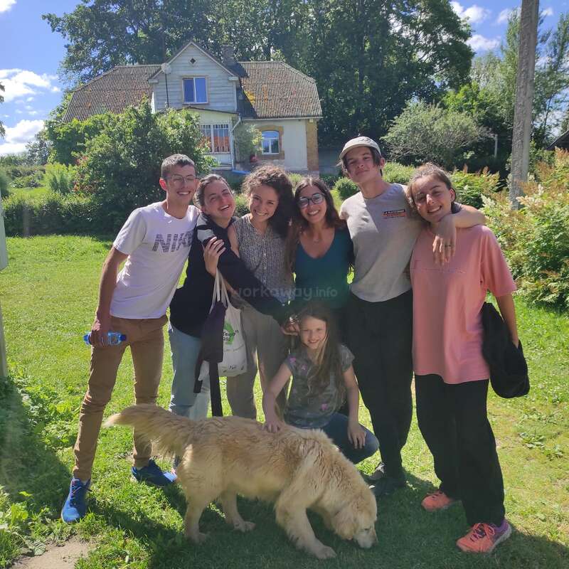 L'image représente un groupe de sept personnes, dont un enfant, posant pour une photo avec un chien devant une maison par une journée ensoleillée, entourée d'une végétation luxuriante.