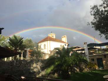 The image depicts a serene scene of a white building with a brown roof, surrounded by lush greenery and a vibrant rainbow stretching across the sky.