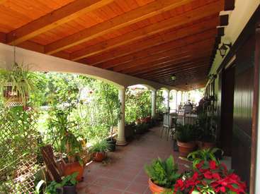 The image depicts a covered patio with a wooden ceiling, white arches, and red tile flooring, surrounded by potted plants and lush greenery.